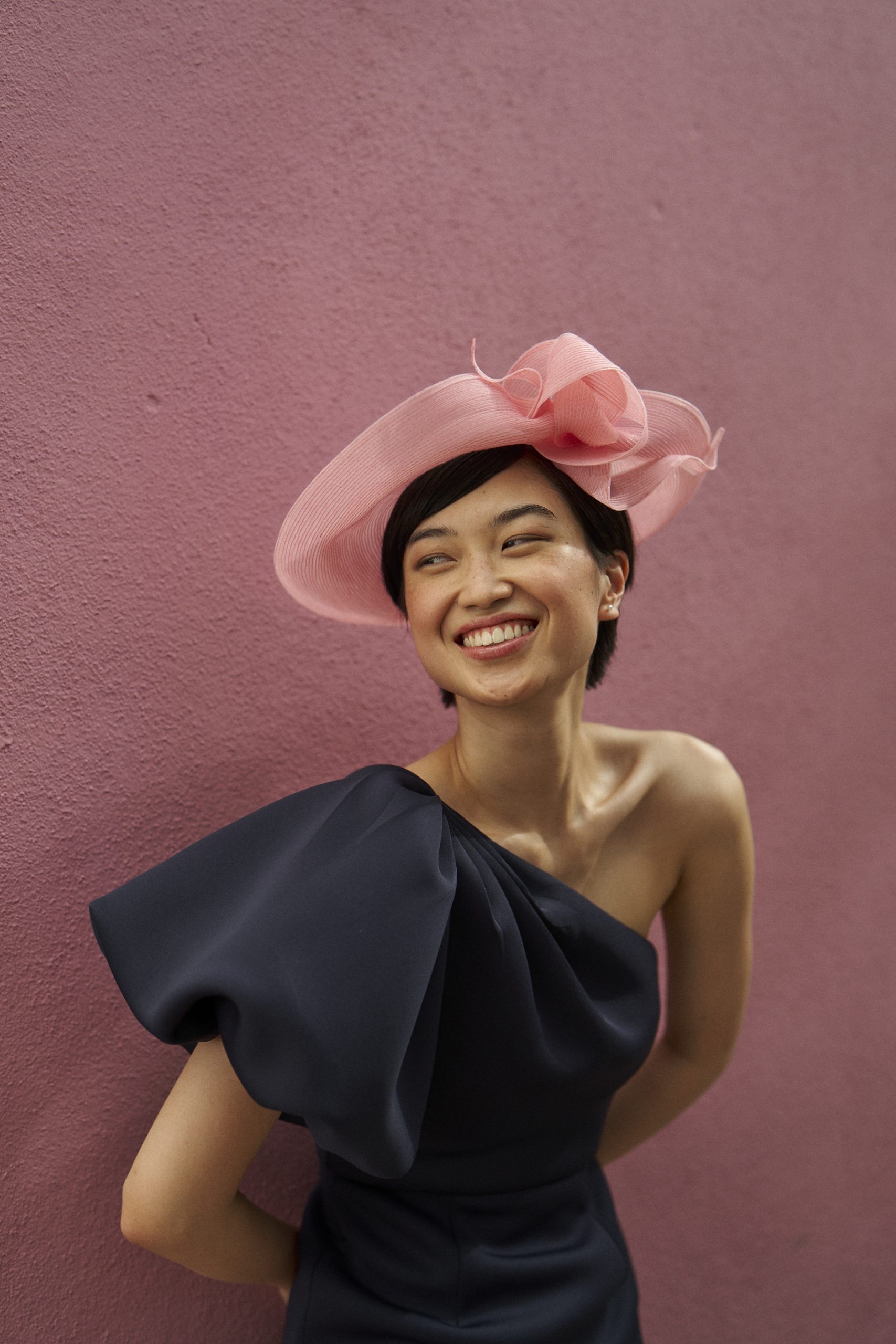 Smiling woman in a large pink hat and dark dress against a pink textured wall.