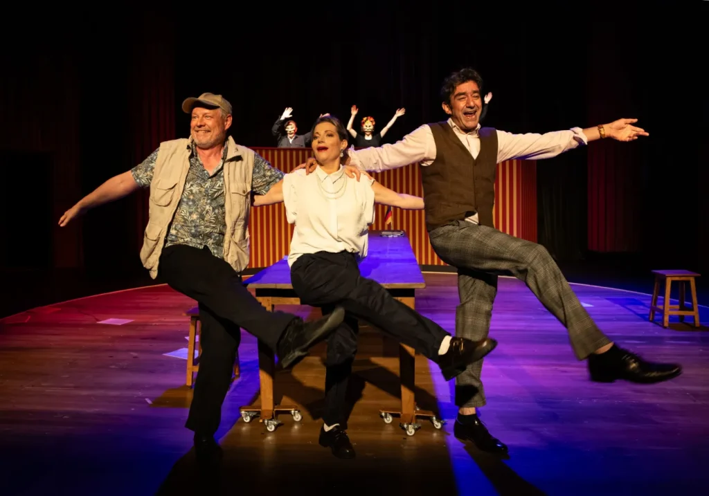 putsch - IN München - Das Stadtmagazin Three actors on a dimly lit stage, arms linked, mid-kick in a playful comedy moment.
