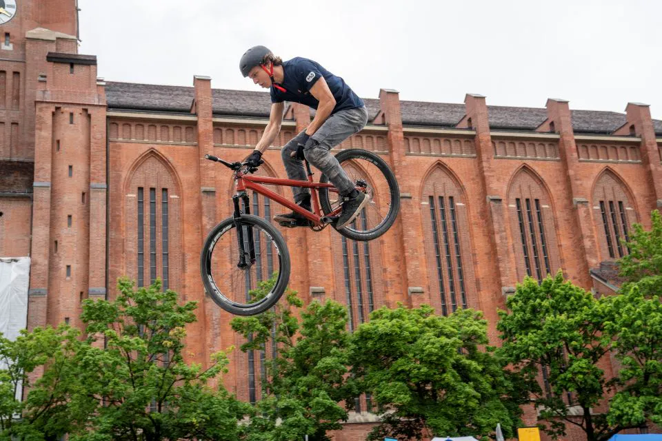 rald-dult-koplett-mor03973 - IN München - Das Stadtmagazin Biker wearing a helmet performs a jump on a red mountain bike against a brick building backdrop.