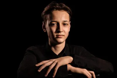 A person in a black shirt sits against a dark background, arms crossed on a surface, looking at the camera with a calm expression.