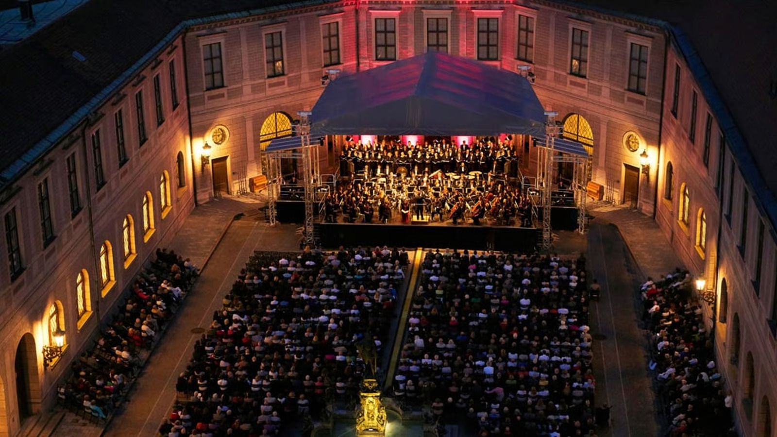 w=1600,h=900,fit=cover (2) - IN München - Das Stadtmagazin Orchestra performing on a stage in an open-air courtyard at night, with audience seated in rows.