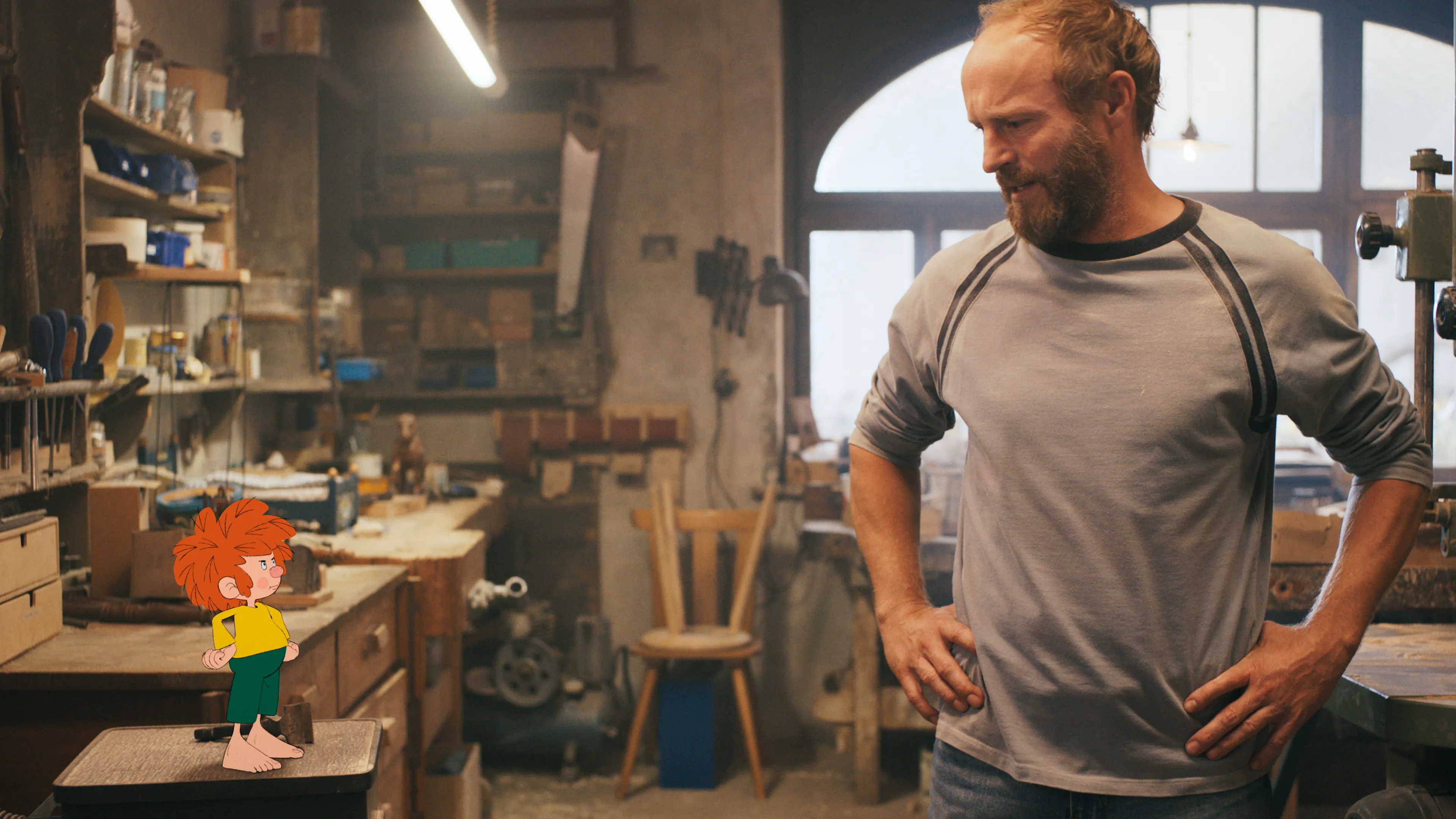 Bearded man in a gray shirt stands in a cluttered workshop, looking down with hands on hips.