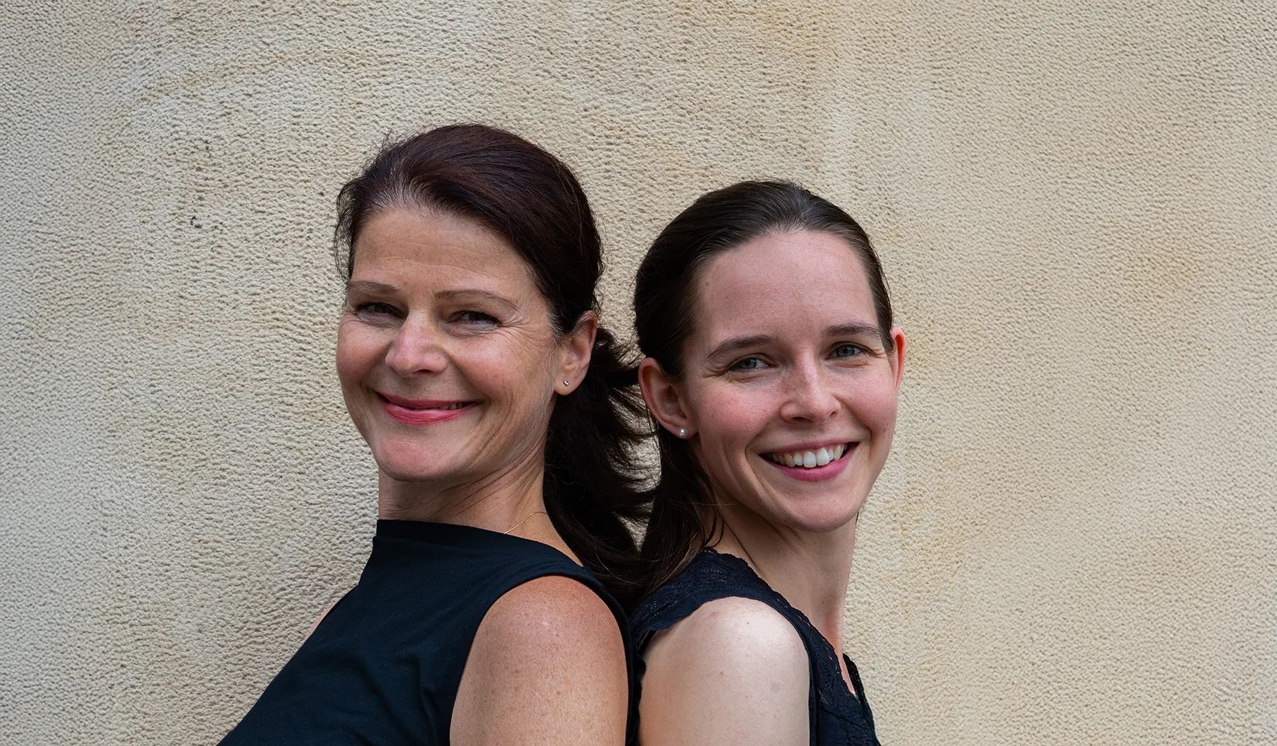 1857x1085-oh-du.schoenes-italien-Unterveranstaltung-Deutsches-Theater-Muenchen-2 - IN München - Das Stadtmagazin Two women smile and stand back-to-back against a beige textured wall.