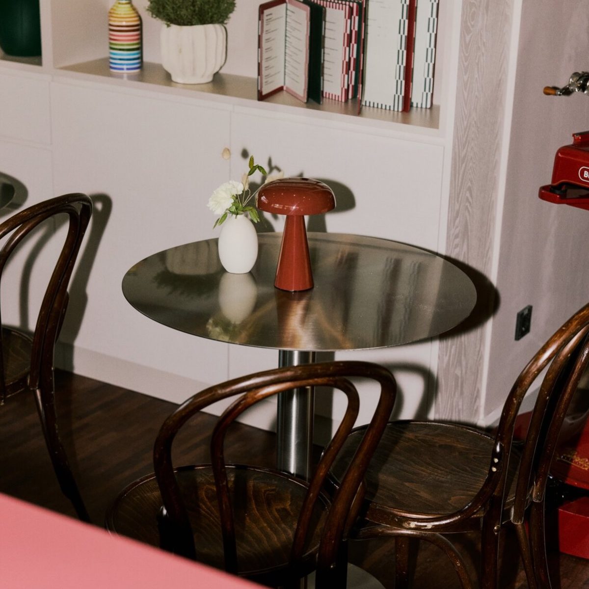 683662134_17877489918590987_5320699370999493127_n - IN München - Das Stadtmagazin Round metal table in a retro cafe setting, featuring a red mushroom-shaped lamp and a small white vase with flowers, surrounded by wooden chairs.