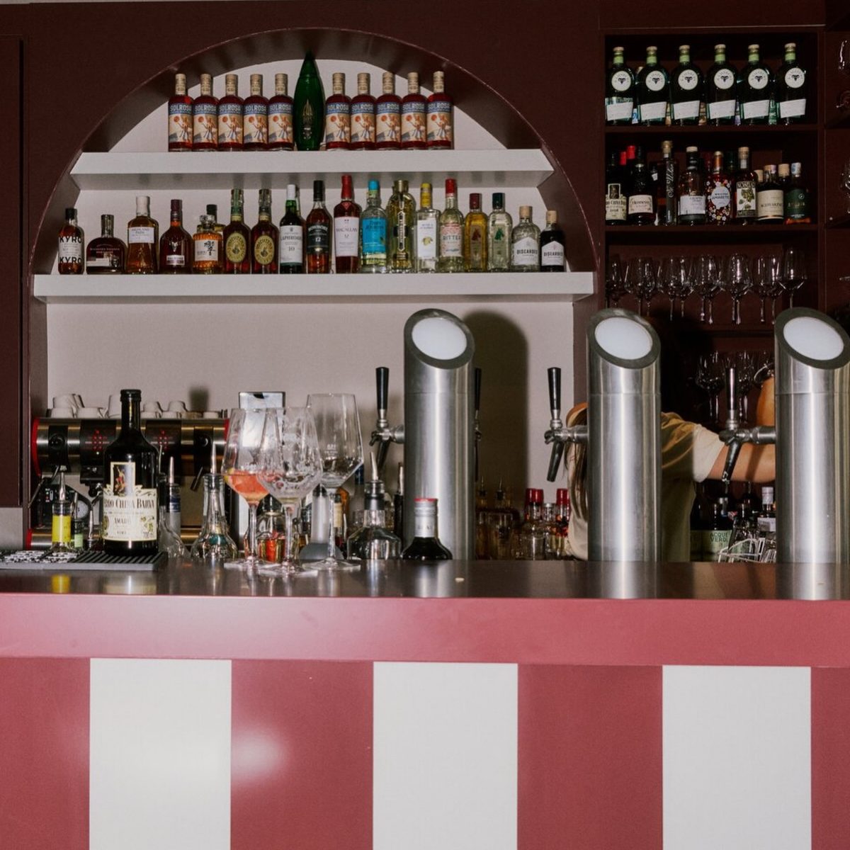 683795205_17877489915590987_2155182106682667368_n - IN München - Das Stadtmagazin Bar counter with red-and-white striped front, liquor shelves behind, and tall beer taps in the foreground.