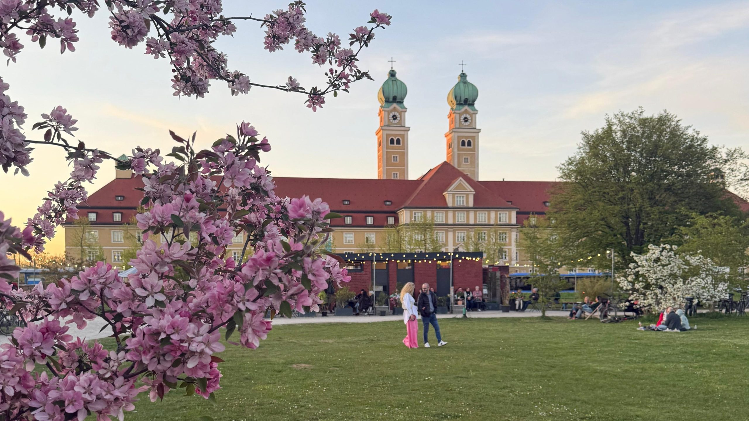 Alexa Steinke - IN München - Das Stadtmagazin Pink blossoms frame a historic building with twin green domed towers in a park at sunset. People mingle near outdoor stalls and string lights.