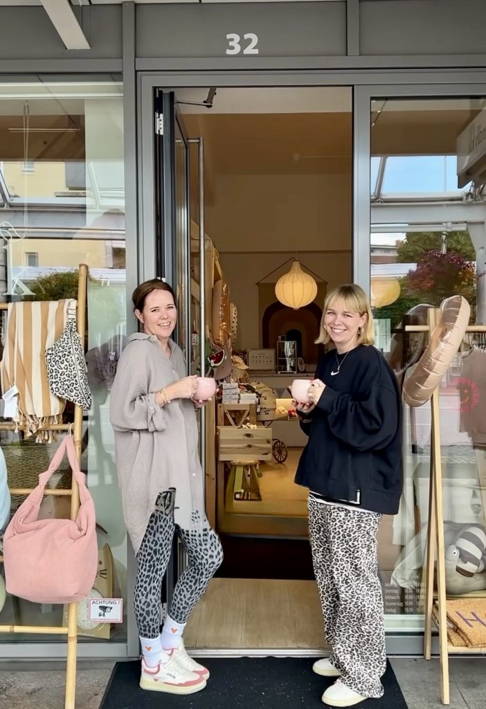 Two women stand at a shop doorway, smiling and holding pink mugs during a sunny day.
