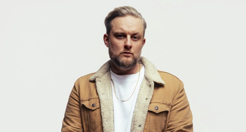 Bildschirmfoto 2026-07-01 um 13.51.42 - IN München - Das Stadtmagazin Portrait of a man with blond hair and a beard wearing a tan shearling-lined jacket, looking at the camera against a light background.