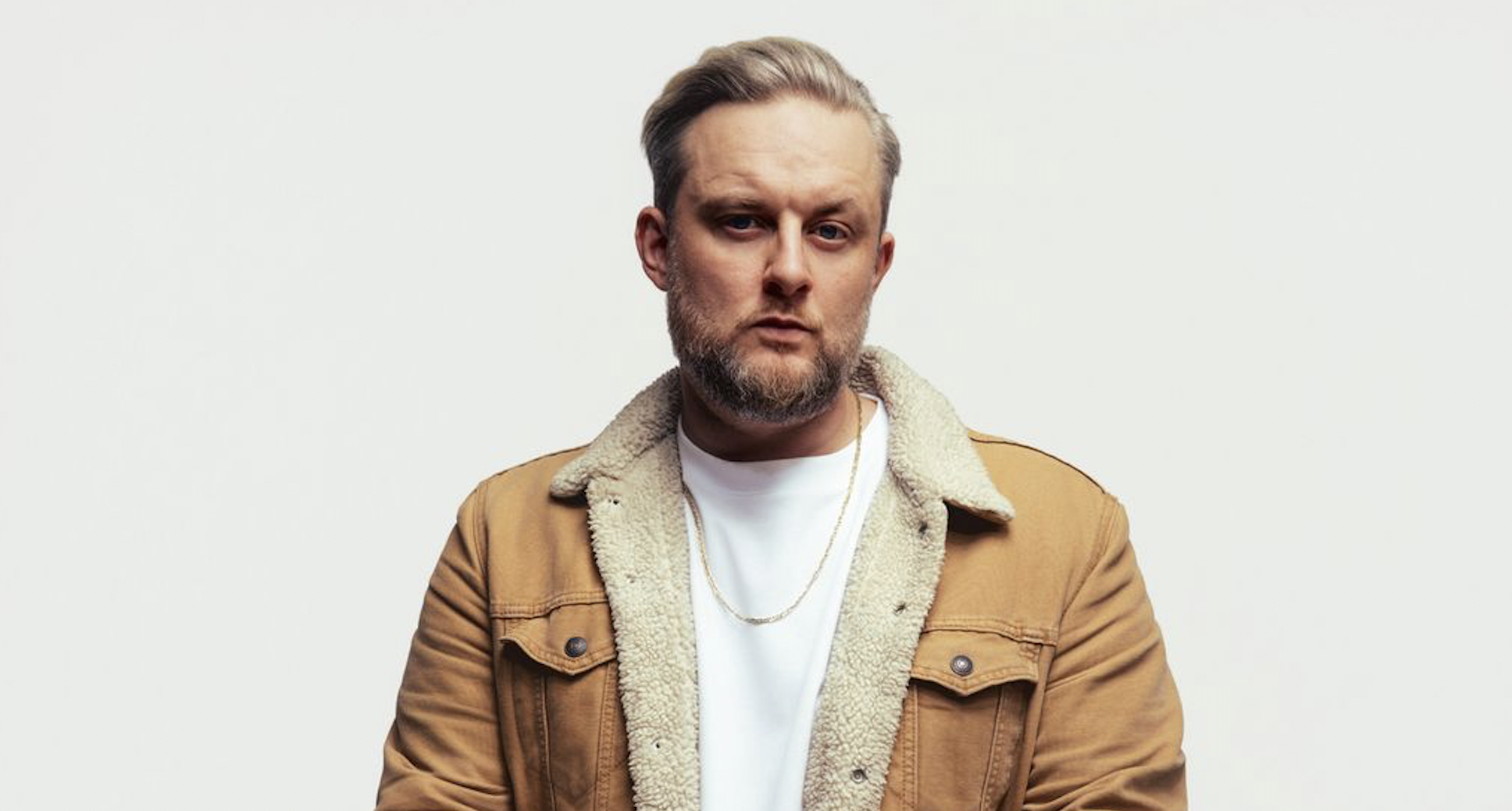 Portrait of a man with blond hair and a beard wearing a tan shearling-lined jacket, looking at the camera against a light background.