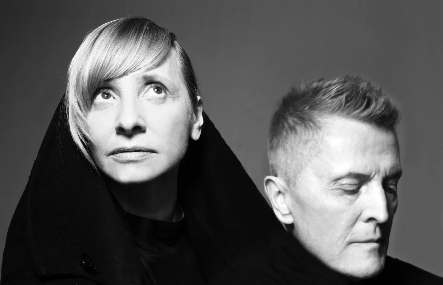 Black-and-white portrait of a woman with short light hair wearing a dark hooded coat, looking upward, beside a man who is looking down.