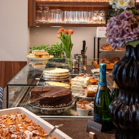 Cafe-Credo-589x882 - IN München - Das Stadtmagazin Glass pastry case with assorted cakes and pastries, a bottle of champagne, and a bouquet on a wooden counter.