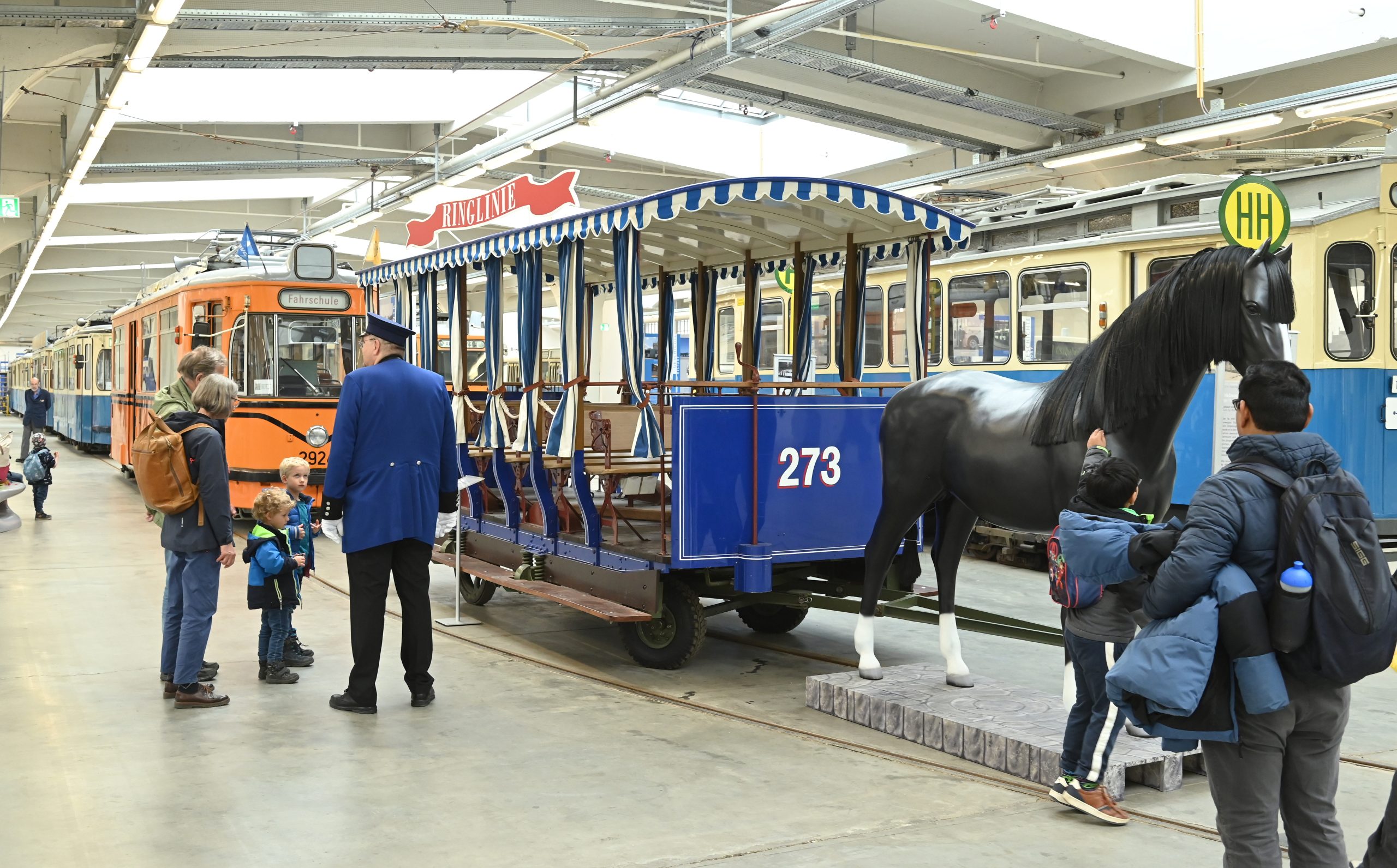 150 Jahre Tram - IN München - Das Stadtmagazin Indoor rail museum showing vintage trams and a blue passenger carriage with striped canopy, visitors watching.