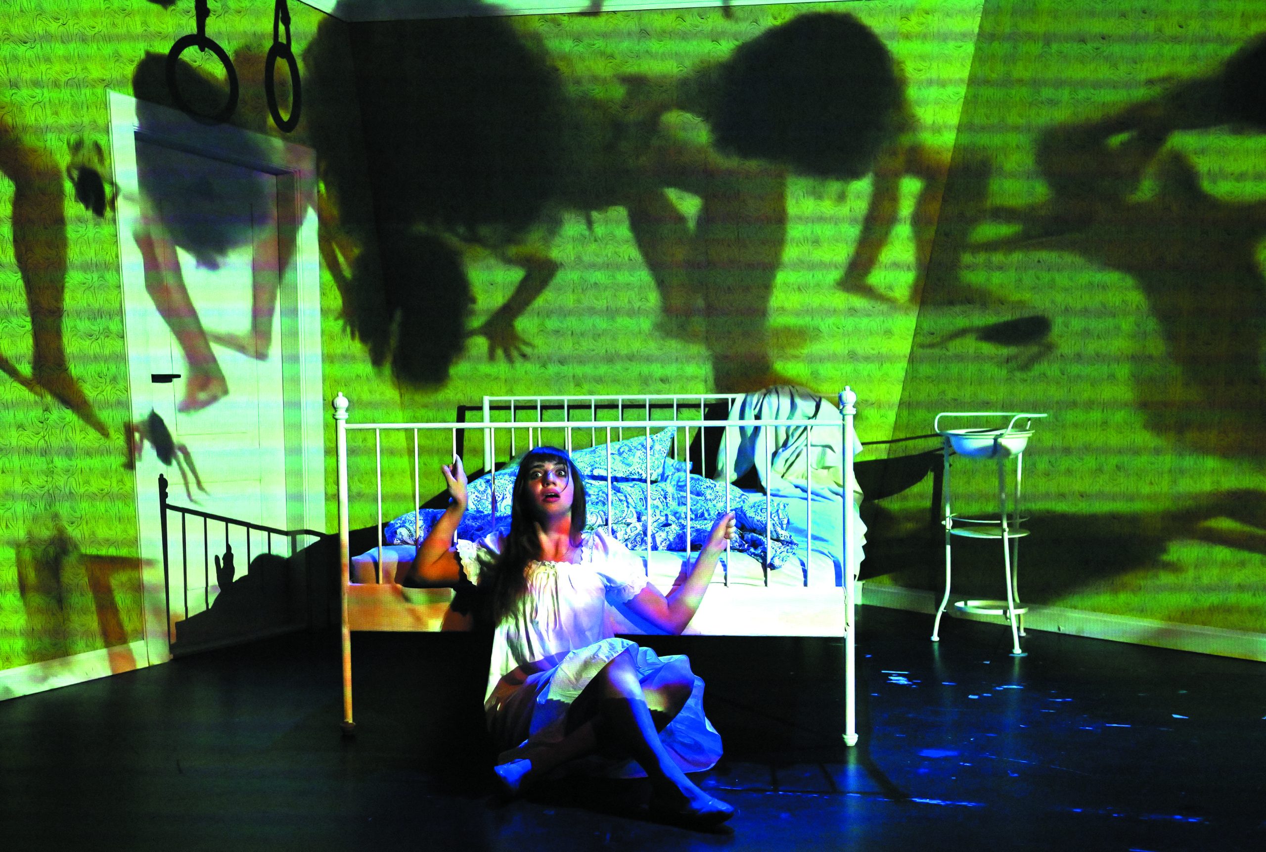 Die gelbe Tapete_45 - IN München - Das Stadtmagazin A girl in a white dress sits on a bed under green lighting, reaching upward as large shadows and silhouettes are projected on a wall behind her, with gym rings hanging nearby.
