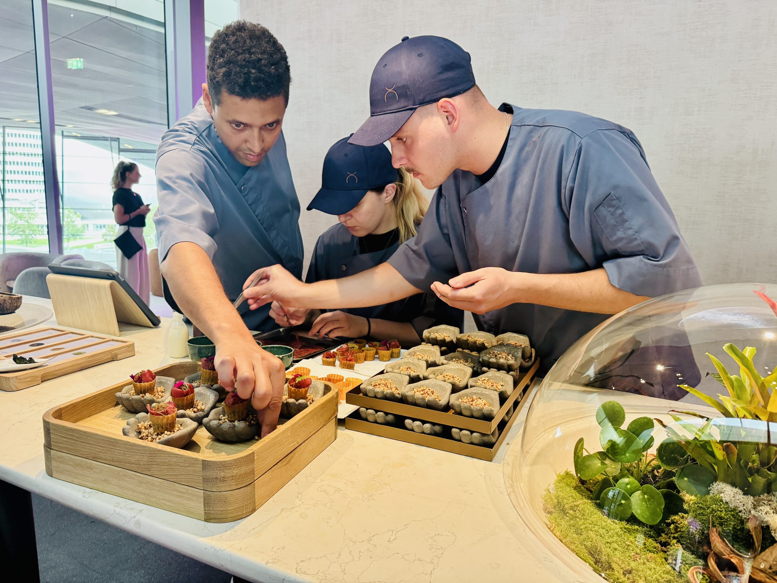 Three chefs in gray uniforms arrange small pastries on wooden trays at a bright food display area.