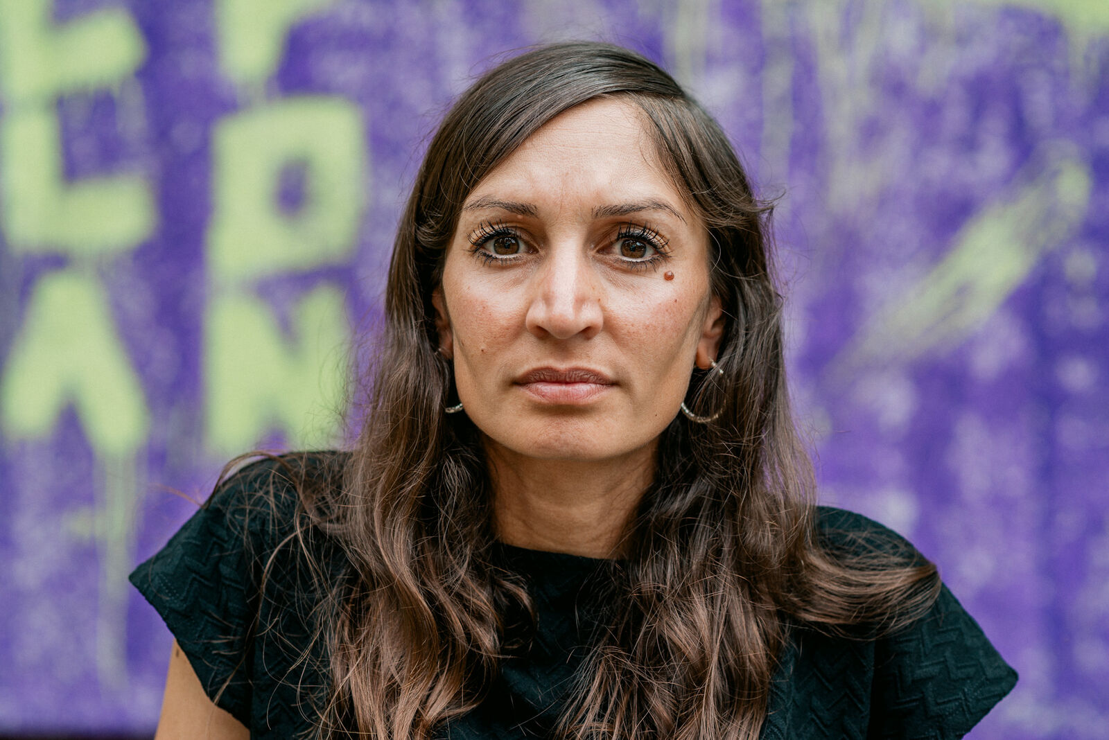 Gilda_Sahebi_c_Hannes_Leitlein - IN München - Das Stadtmagazin Portrait of a woman with long brown hair and hoop earrings, looking directly at the camera, against a purple graffiti wall.