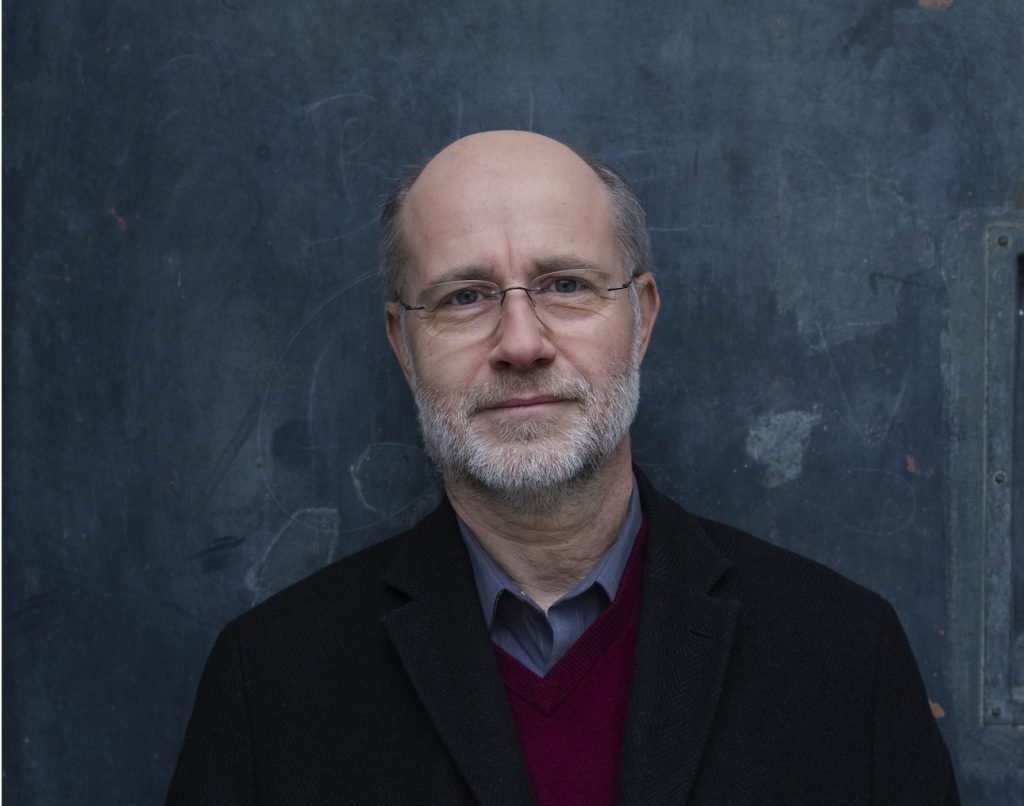 Harald Lesch - IN München - Das Stadtmagazin Portrait of a middle-aged man with glasses and a gray beard, wearing a dark blazer against a blue textured wall.