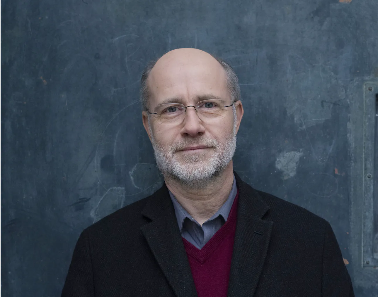 Portrait of a middle-aged man with glasses and a gray beard, wearing a dark blazer against a blue textured wall.