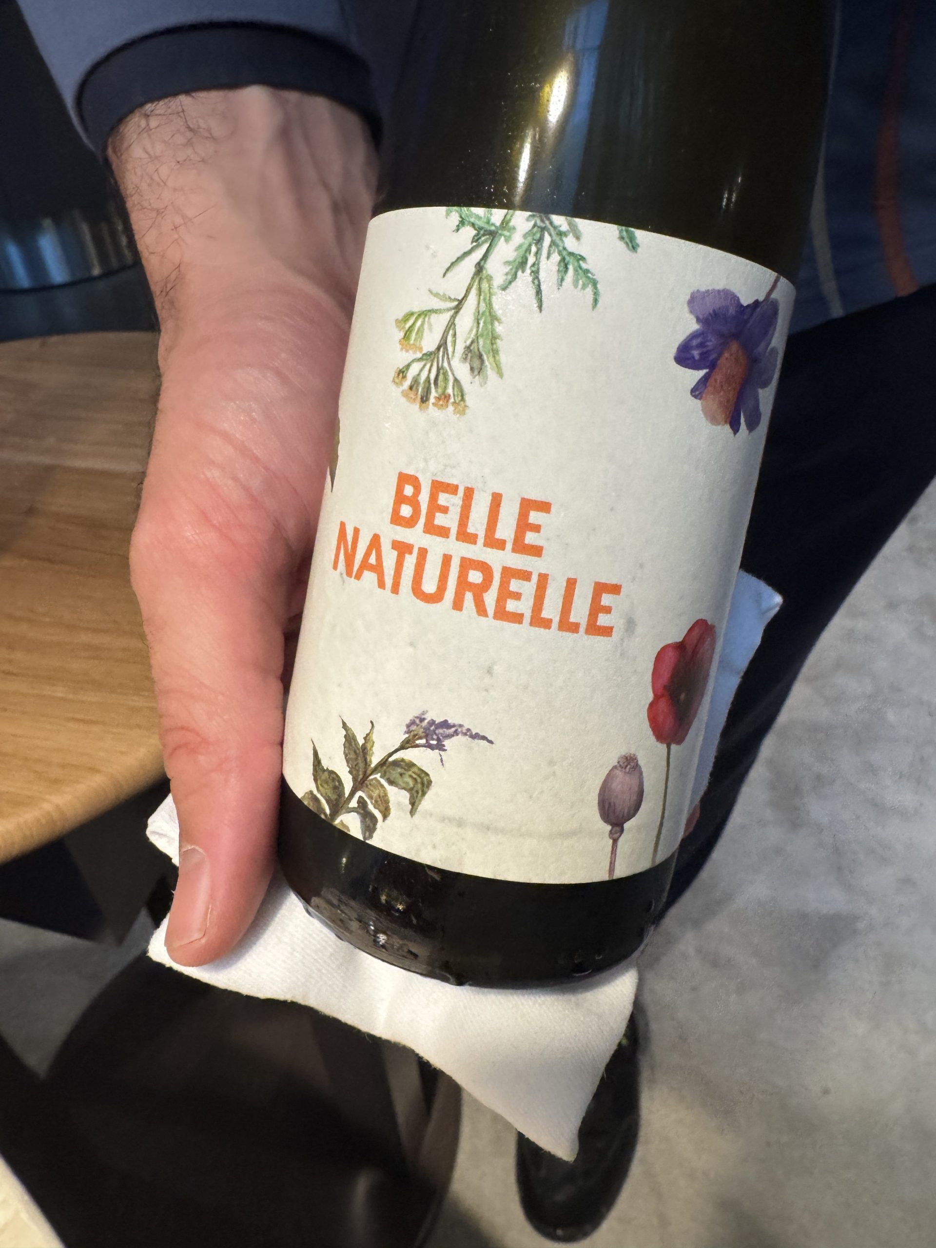IMG_9678 - IN München - Das Stadtmagazin Hand holding a wine bottle with a floral label reading 'BELLE NATURELLE' in orange letters.