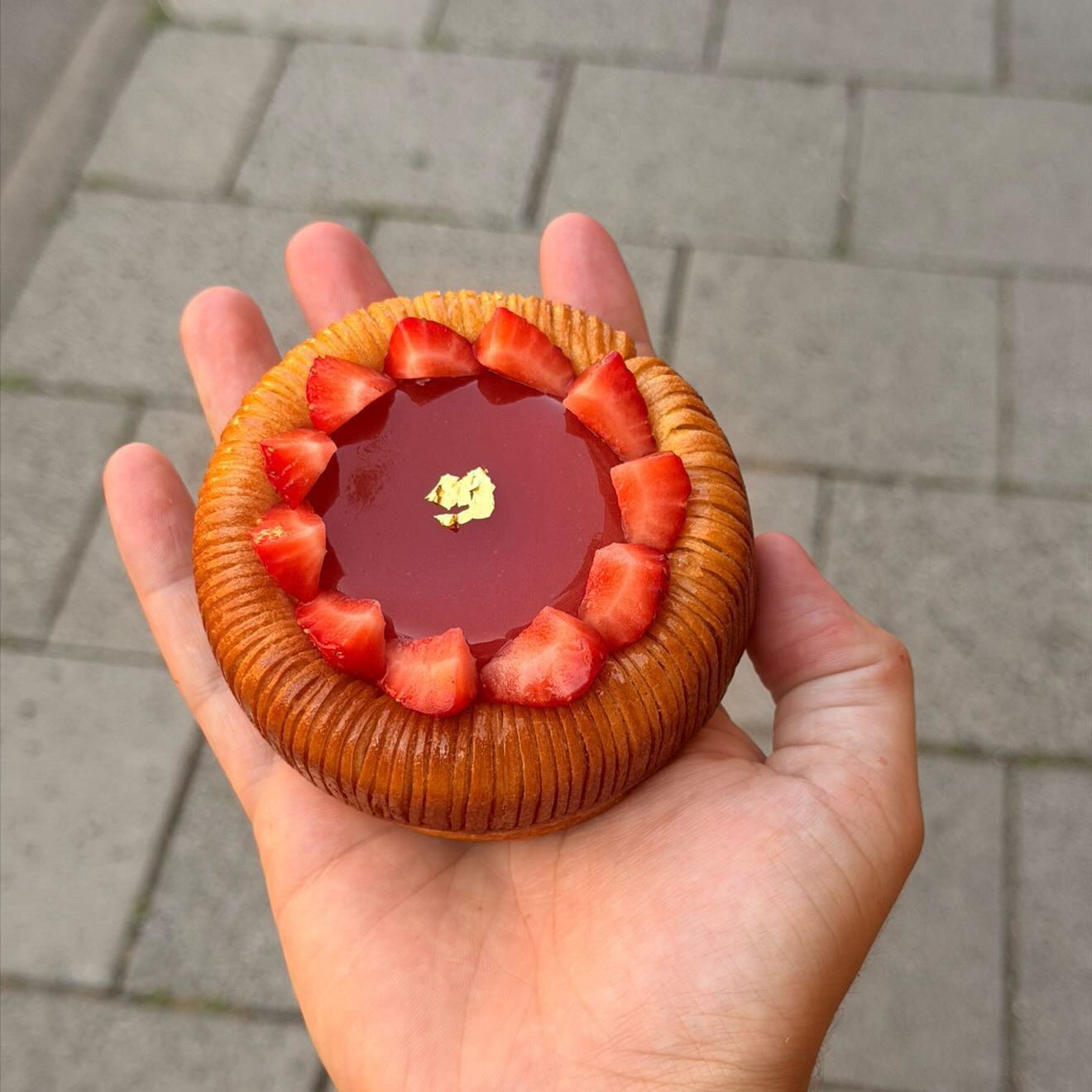 Jamie Sun_2 - IN München - Das Stadtmagazin Round tart held in a palm, with sliced strawberries around the edge and a glossy red filling topped with gold leaf, outdoors on paving stones.