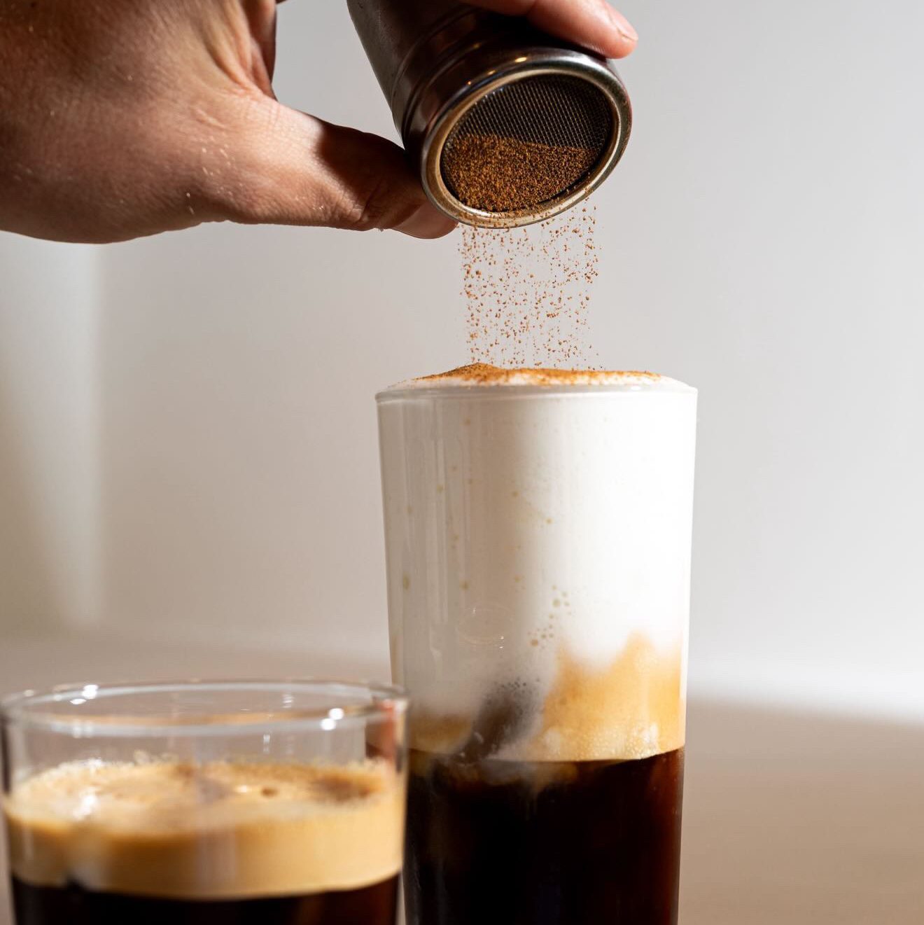 Jamie Sun_3 - IN München - Das Stadtmagazin Hand pouring ground coffee from a shaker into a tall glass of dark espresso, with crema forming on top.