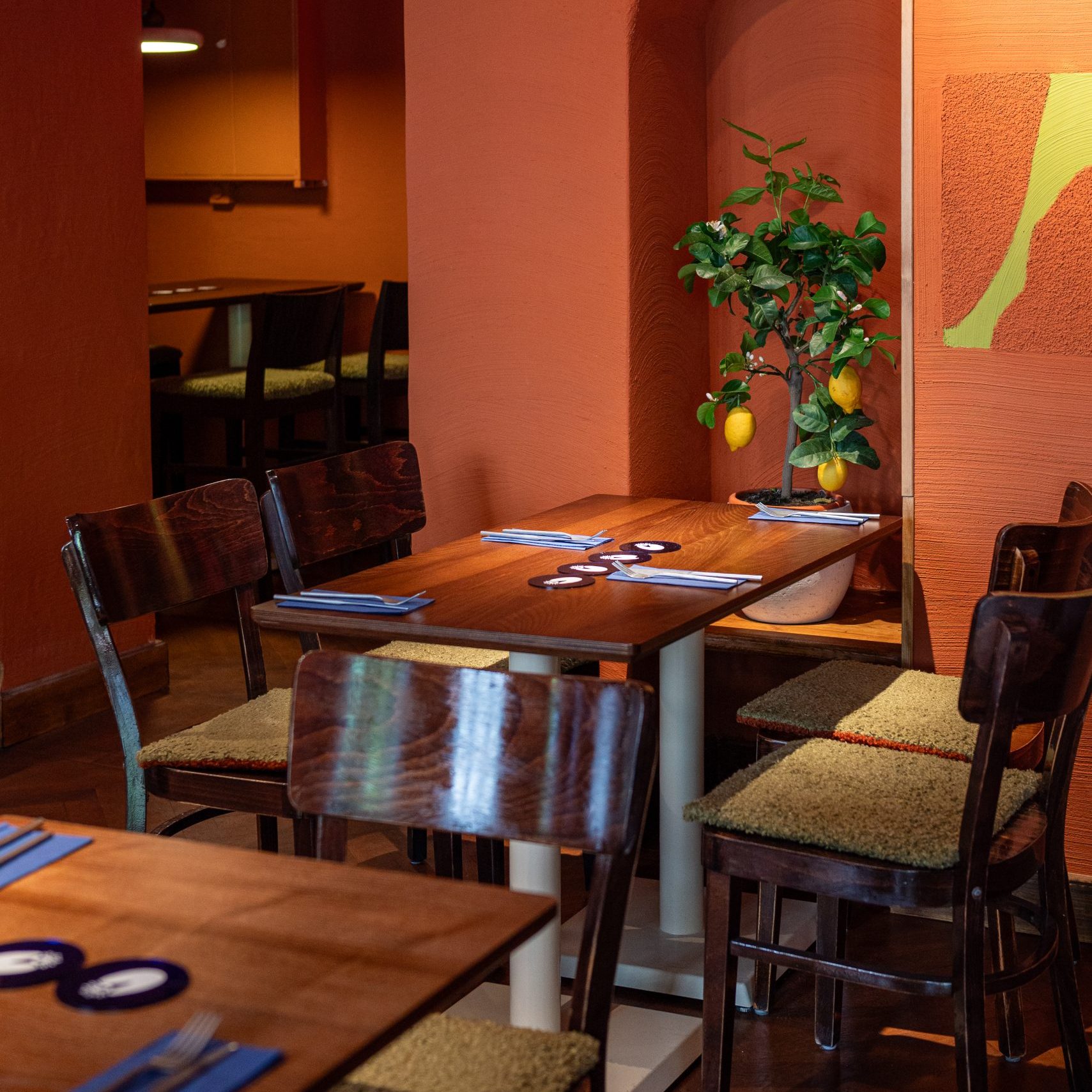 Janina Amendt_2 - IN München - Das Stadtmagazin Cozy dining area with orange walls, wooden tables and chairs; a potted lemon tree sits by the wall.