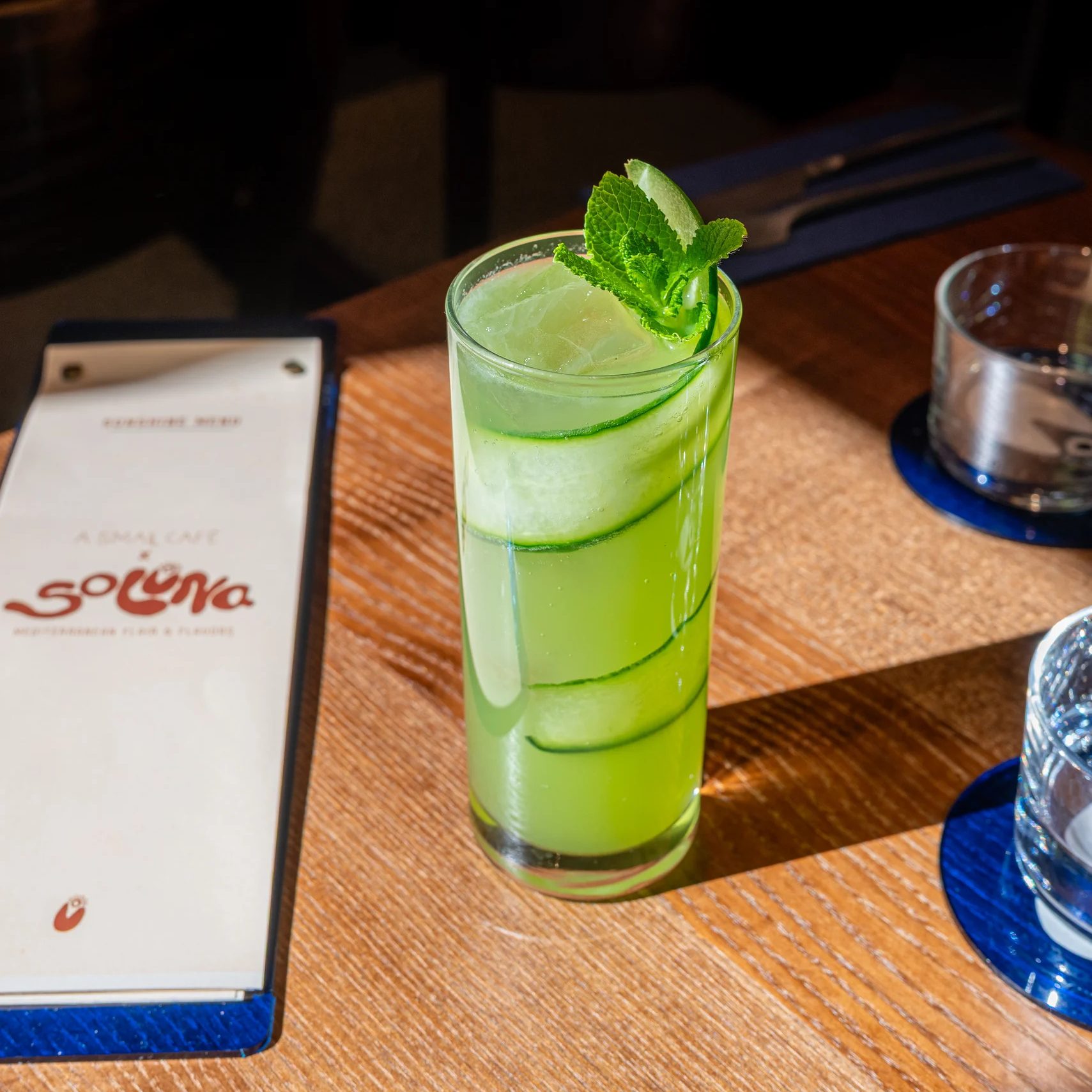 Janina Amendt_4 - IN München - Das Stadtmagazin Tall green mojito with mint garnish on a wooden table, beside a menu and blue coasters.