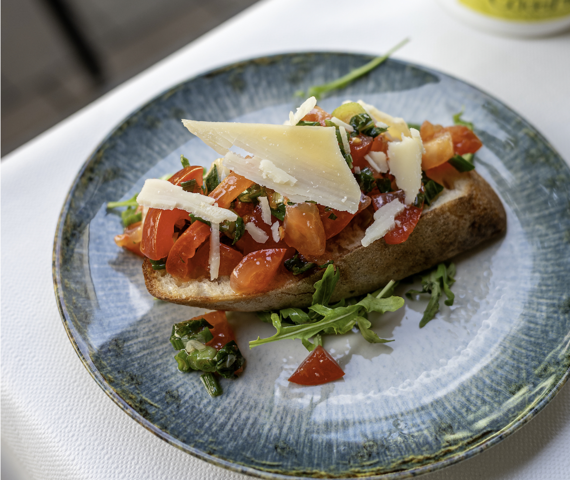 Crusty bread slice topped with tomato mixture, arugula, basil and shaved Parmesan on a blue plate.