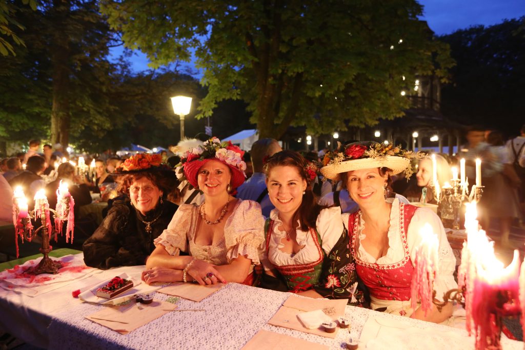 Kocherlball München - IN München - Das Stadtmagazin Group of smiling women in traditional folk dresses with flowered hats sit at a candlelit outdoor table during a festive evening event.