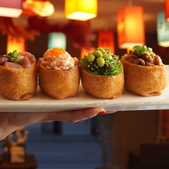 Komo - IN München - Das Stadtmagazin Four inari sushi bites on a plate held by a hand, each topped with different fillings (tuna, spicy tuna, greens with edamame, sesame) with colorful lanterns in the background.