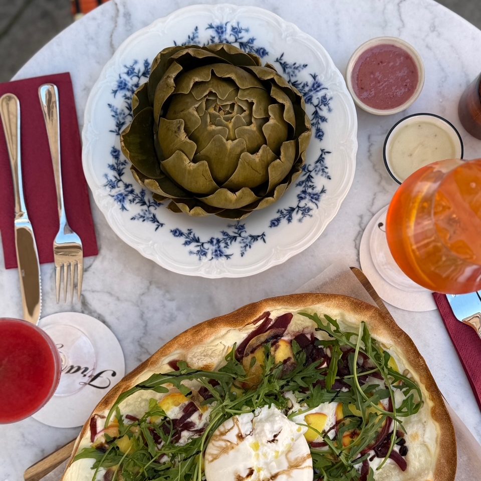 Martina Grandjean_3 - IN München - Das Stadtmagazin Pizza topped with burrata, arugula, and colorful vegetables on a round pizza at an outdoor table with drinks nearby.