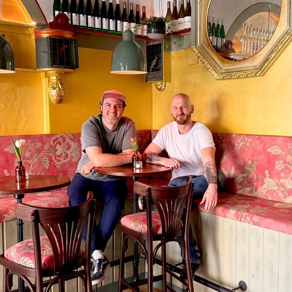 Martina Grandjean_4 - IN München - Das Stadtmagazin Two men sit at a round table in a colorful cafe, pink floral booth seating, yellow walls, and bottles lined on a shelf behind them.