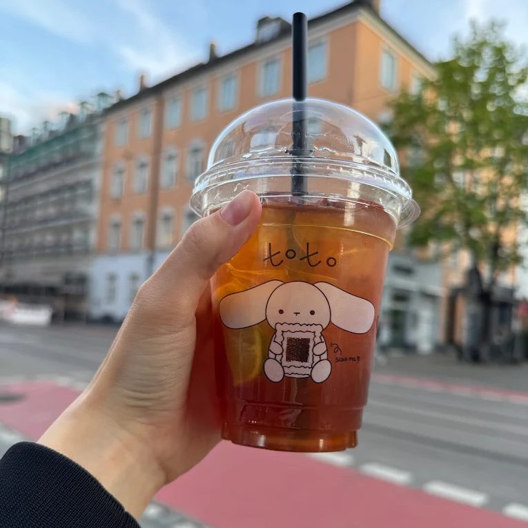 Mary Pergunova - IN München - Das Stadtmagazin Hand holding iced coffee in a clear cup with dome lid and black straw, featuring a cute dog drawing and 'to.to' logo, outdoors on a city street.