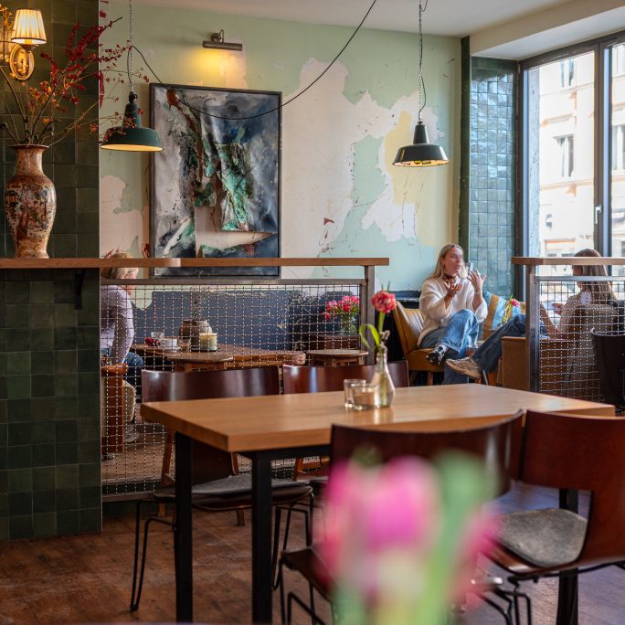 Mirin-3-684x1024 - IN München - Das Stadtmagazin Cozy café interior with wooden tables, green-tiled column, wall art, and two women talking by a large window.