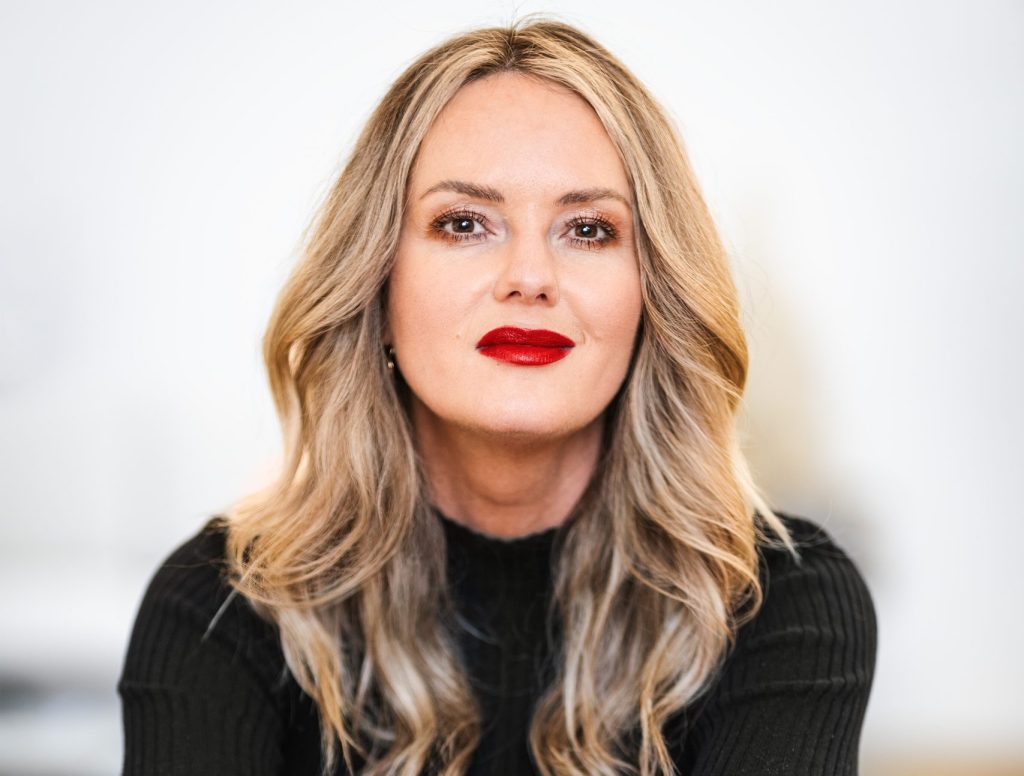 Mona Fuchs - IN München - Das Stadtmagazin Close-up of a woman with long blonde hair and red lipstick, wearing a black top, facing the camera with a neutral background