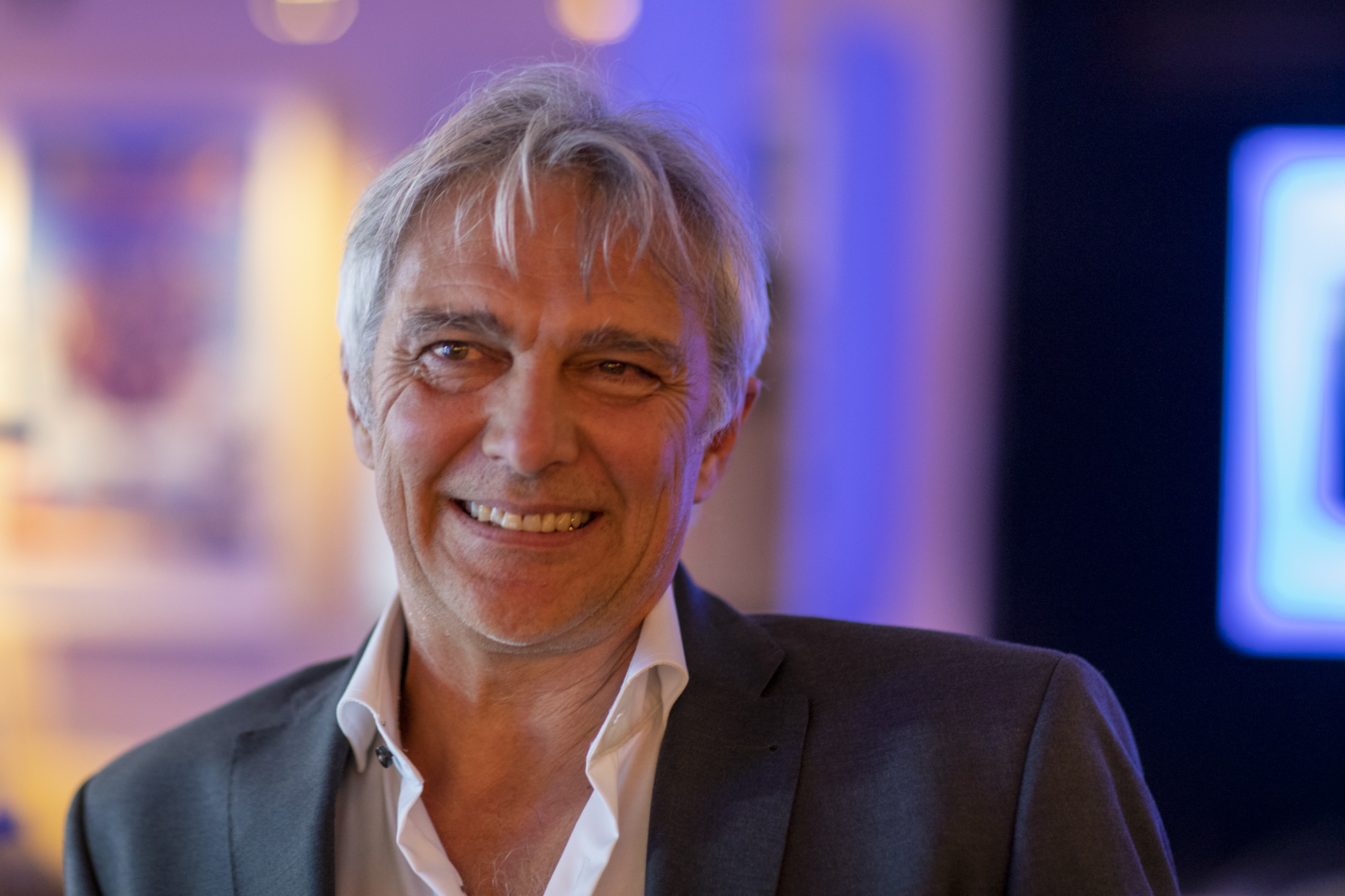 Smiling older man in a dark suit jacket and white shirt, colorful blurred background at a social event.