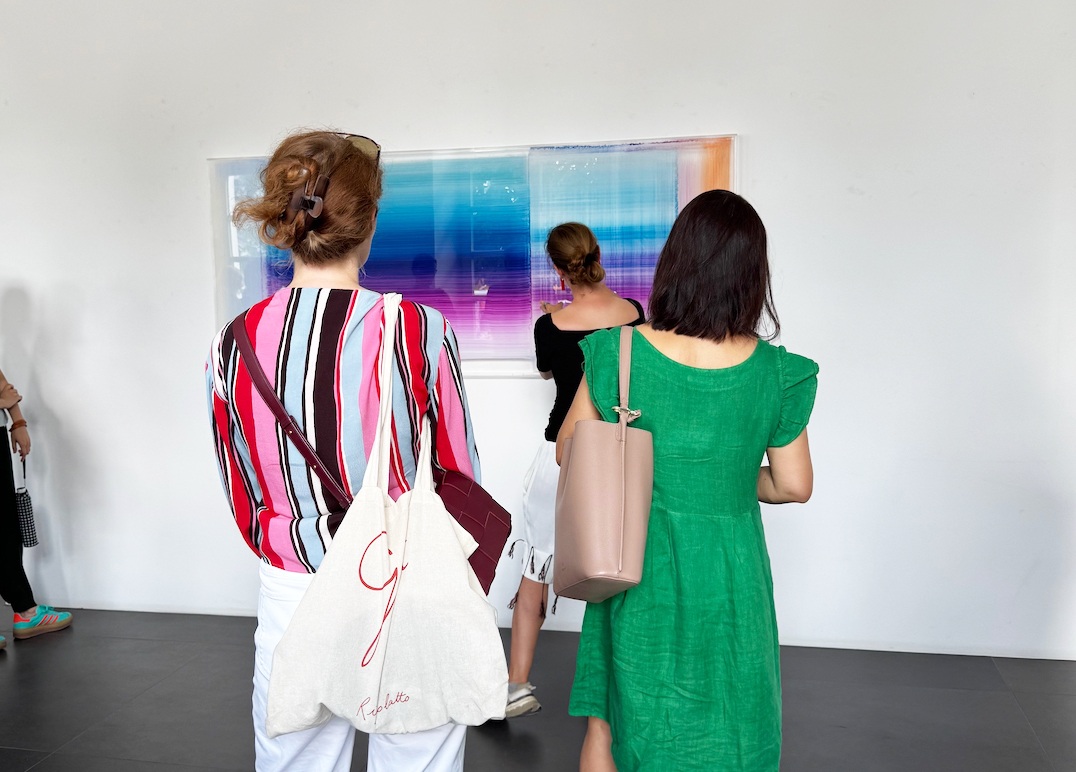 People in a bright gallery viewing an abstract gradient artwork with blue to orange hues on the wall, carrying bags and wearing colorful clothing.
