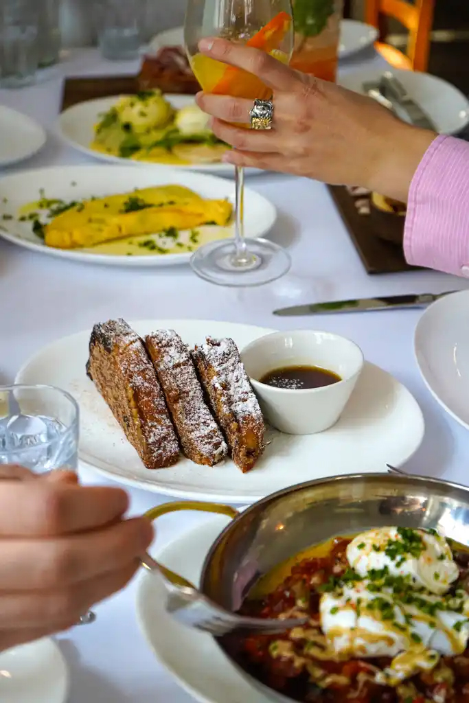 Open_Art_Preysinggarten_Brunch1 - IN München - Das Stadtmagazin Person toasting with a wine glass over a table of plated brunch dishes; foreground shows powdered-sugar churros with dip.