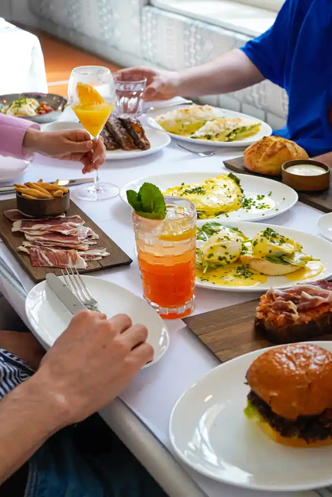Open_Art_Preysinggarten_Brunch2 - IN München - Das Stadtmagazin People dining at a table with a variety of dishes: burgers, charcuterie, eggs, tapas, bread, and cocktails being shared.