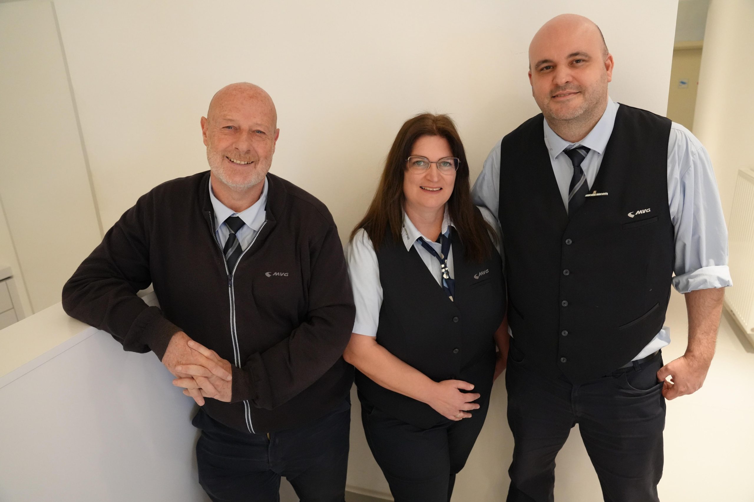 Sprecher-Texter-MVG_Audio_Guide - IN München - Das Stadtmagazin Three staff members in uniforms pose together indoors, leaning against a white wall and smiling (two men in vests, woman in center).