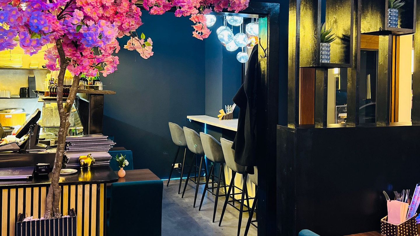 Thanh Huynh_1 - IN München - Das Stadtmagazin Indoor cafe/bar area with a pink flowering tree beside a dark blue wall and a row of beige bar stools along a high counter under glass globe lights.
