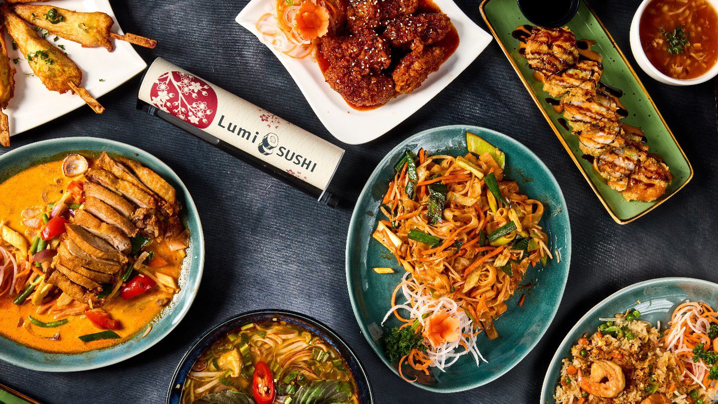 Thanh Huynh_2 - IN München - Das Stadtmagazin Assorted Asian dishes on a dark table: curry, noodles, sushi, and fried items plate-trio with Lumi Sushi bottle in center