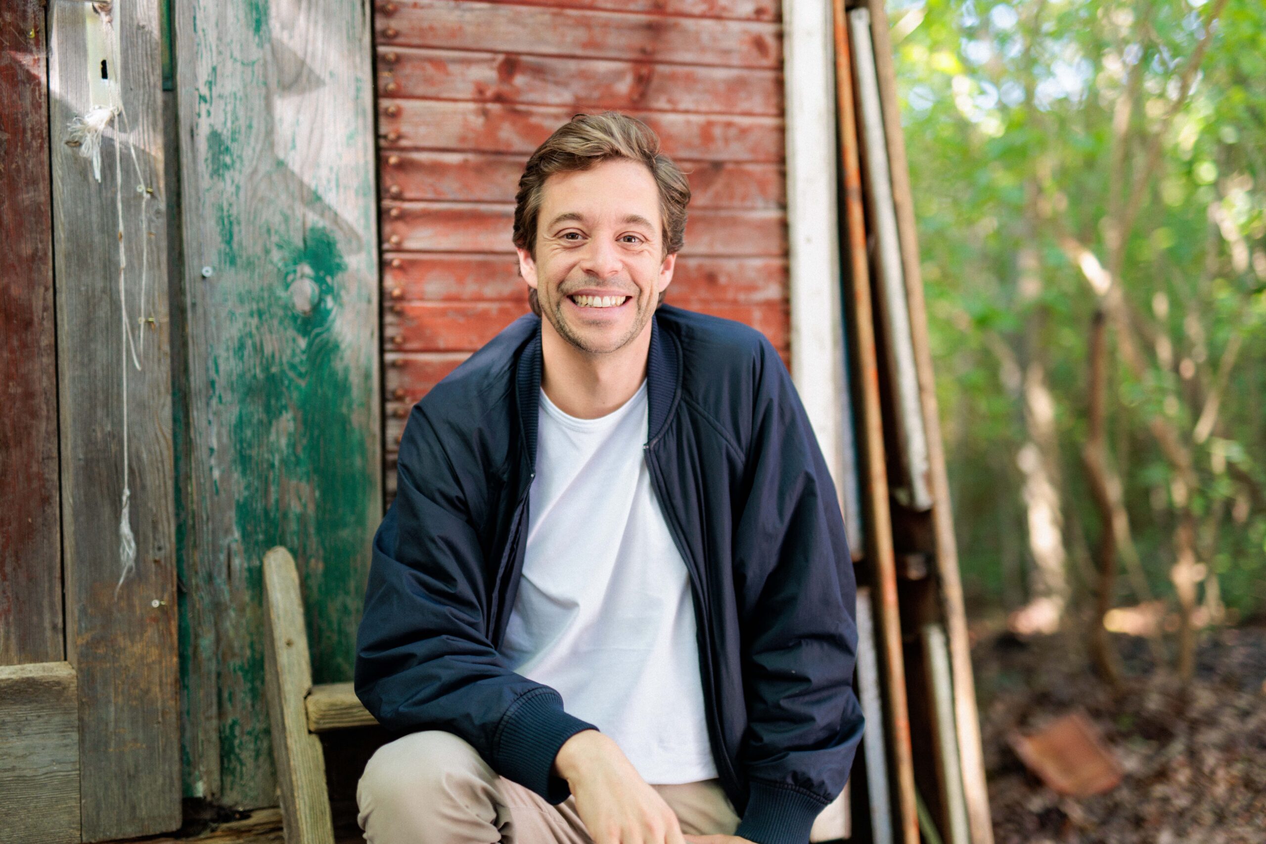 TobiPortraits_Sept24_HFH-36-Kopie-scaled - IN München - Das Stadtmagazin Young man sitting on weathered wooden steps beside a rustic shed, smiling at the camera.