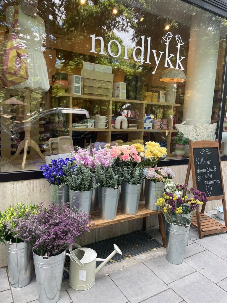 Storefront display with metal buckets of colorful flowers outside a shop window and a chalkboard sign on the right.