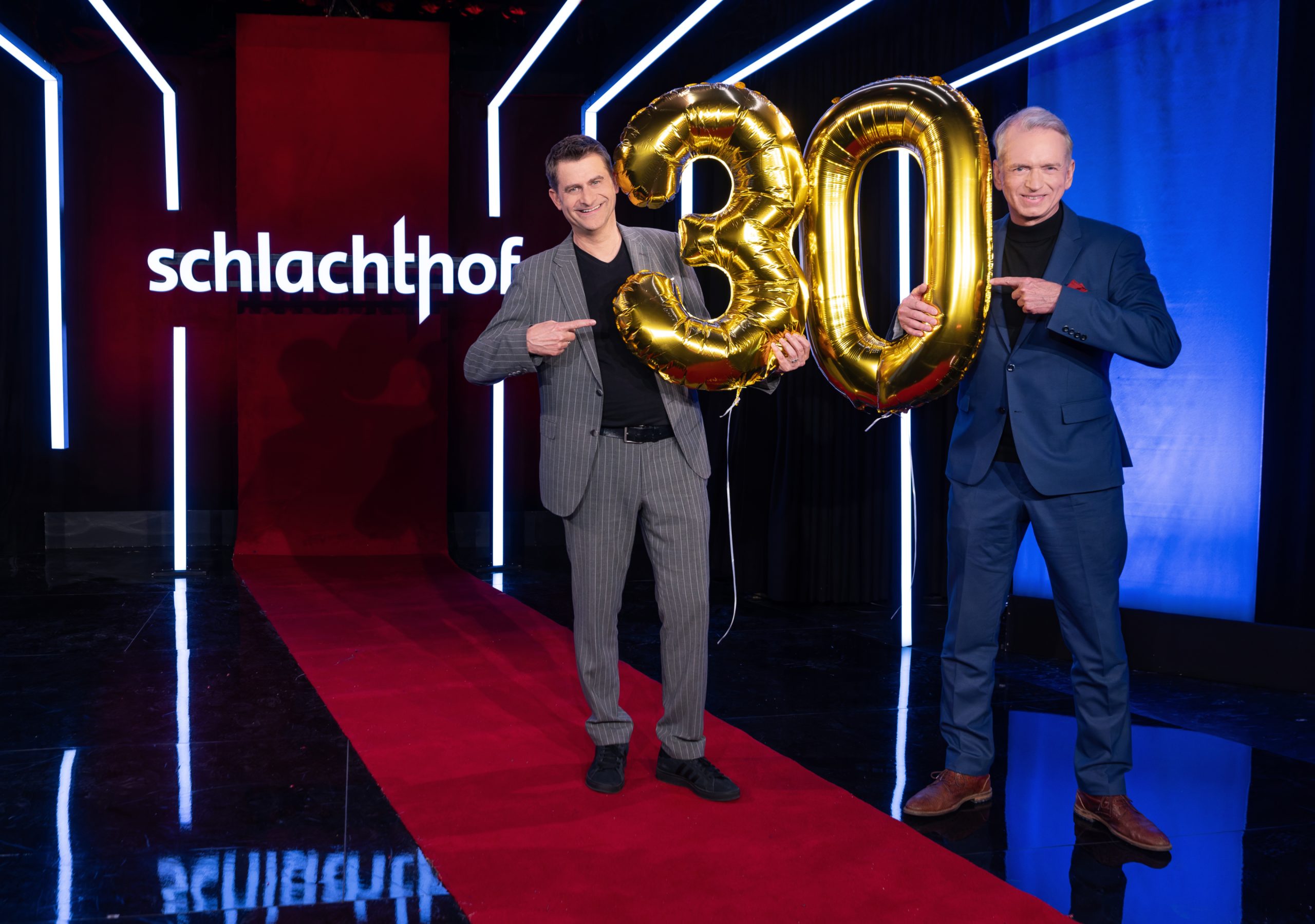 Two men in suits pose on a red carpet, holding gold '30' balloons in a neon-lit venue with the Schlachthof sign visible in the background.
