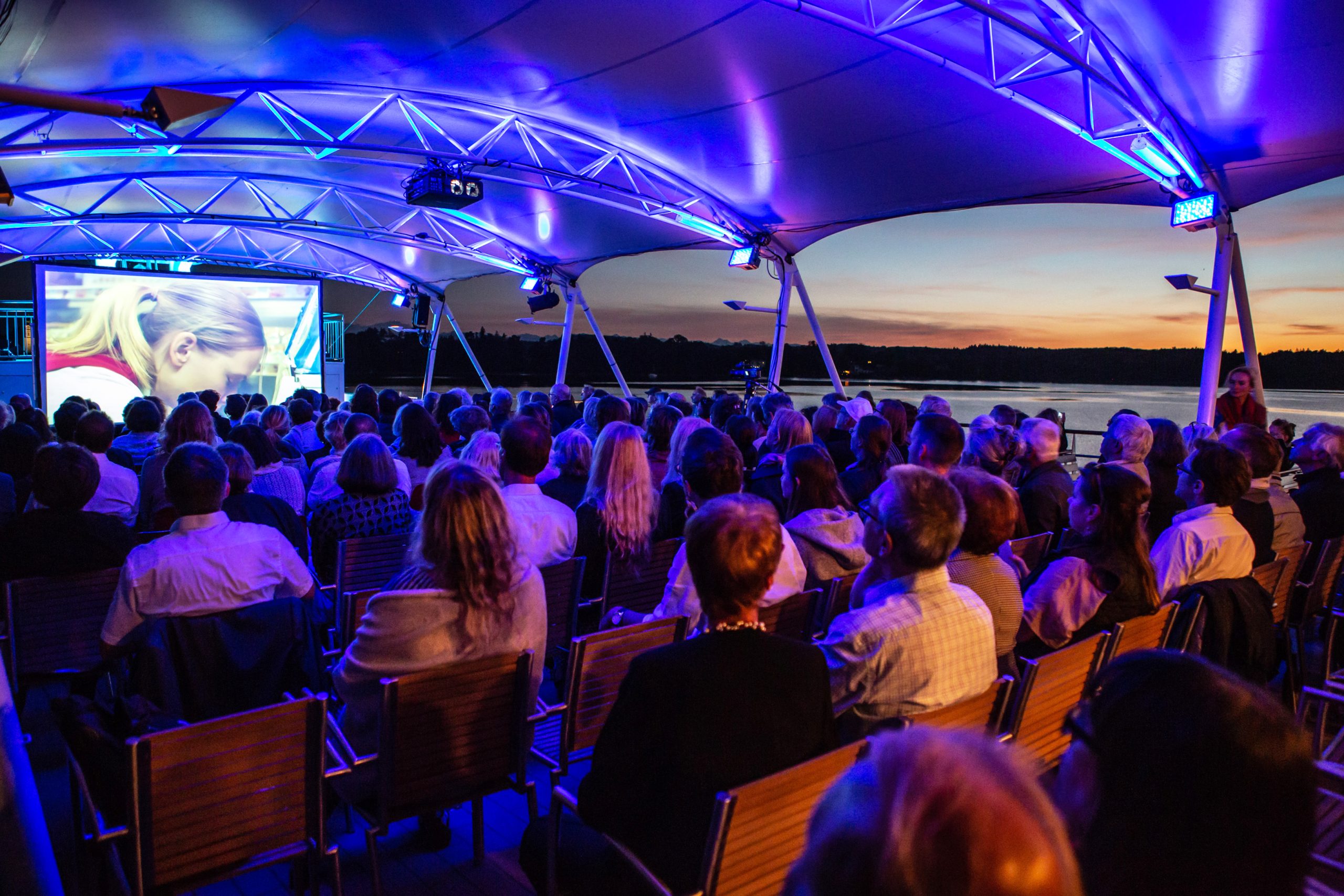 Large audience seated in an outdoor riverside venue under blue lighting, watching a screen with a person on it as sunset colors glow over the water