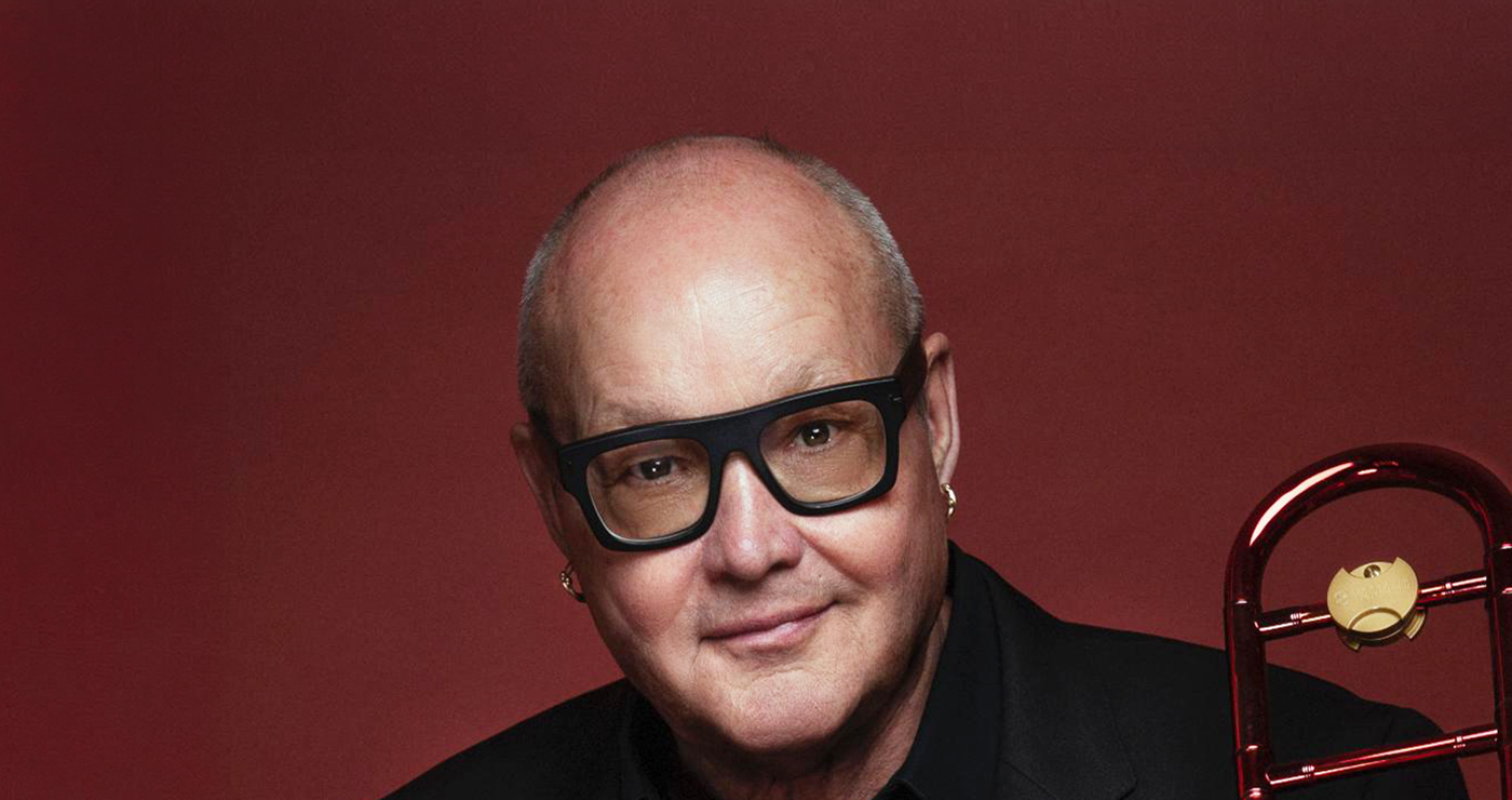 Smiling man with large black-framed glasses in a black suit, red studio backdrop, with a brass trombone visible on the right side.