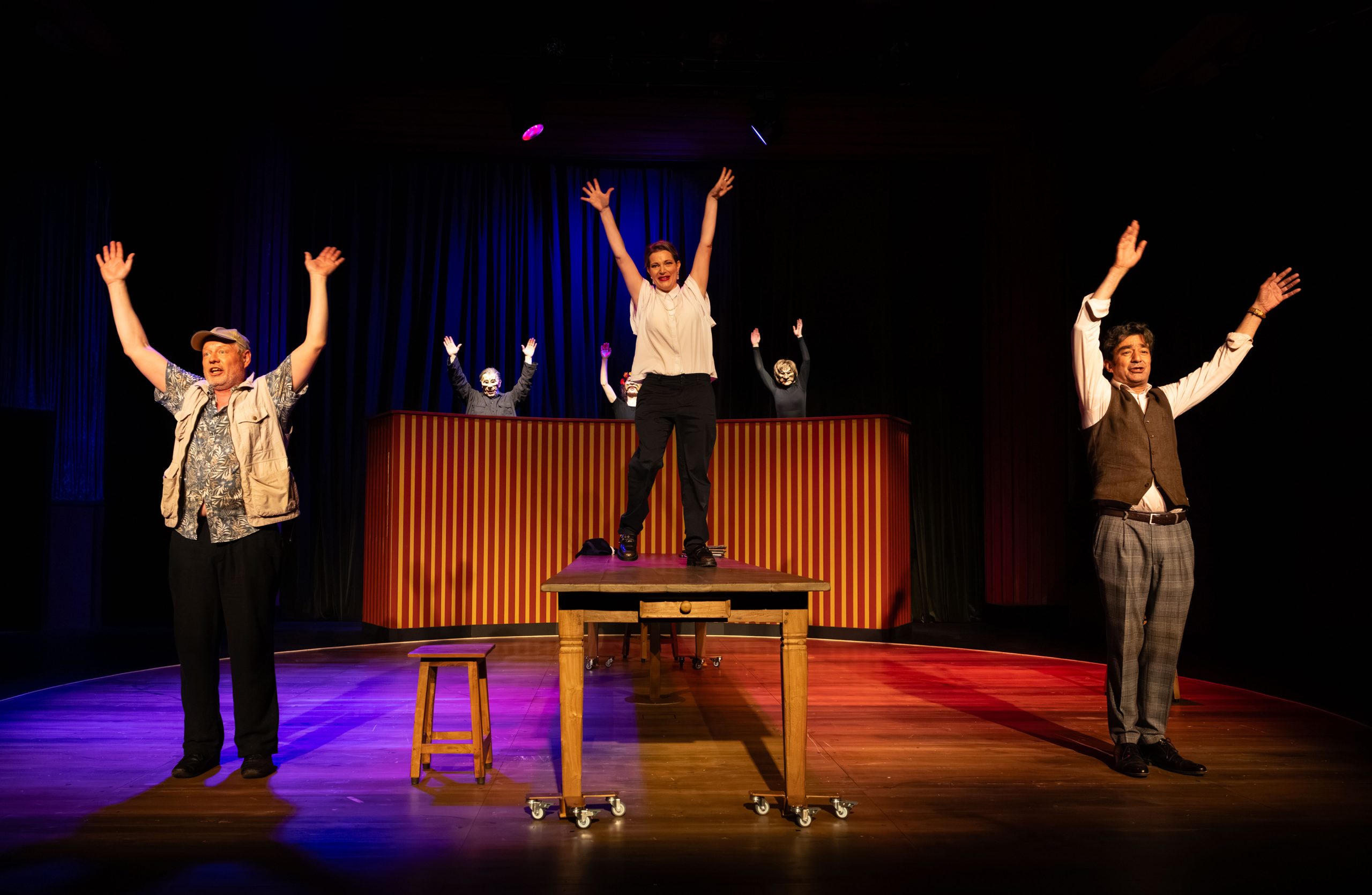 Woman center stage on a raised platform with arms raised, flanked by performers on either side in a theater with striped backdrop and blue lighting.