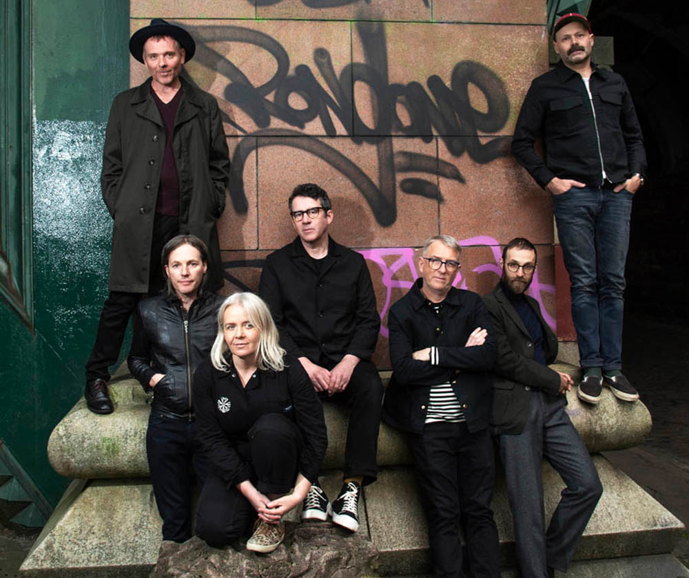 Seven people—a band—posing together on a stone bench in an urban, graffiti-covered setting, with most wearing dark jackets and casual shoes.