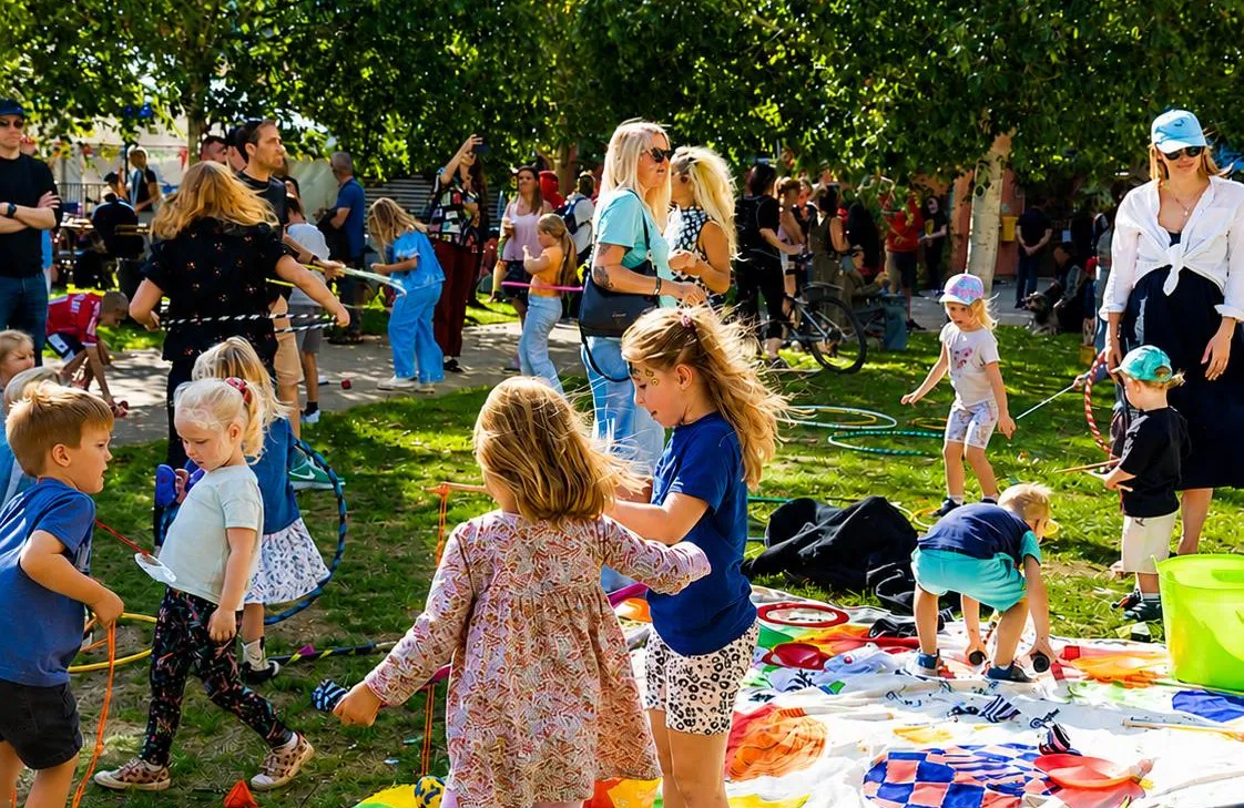Kids and adults enjoy a sunny park gathering; children play with hoops and blankets while caregivers supervise in the background.