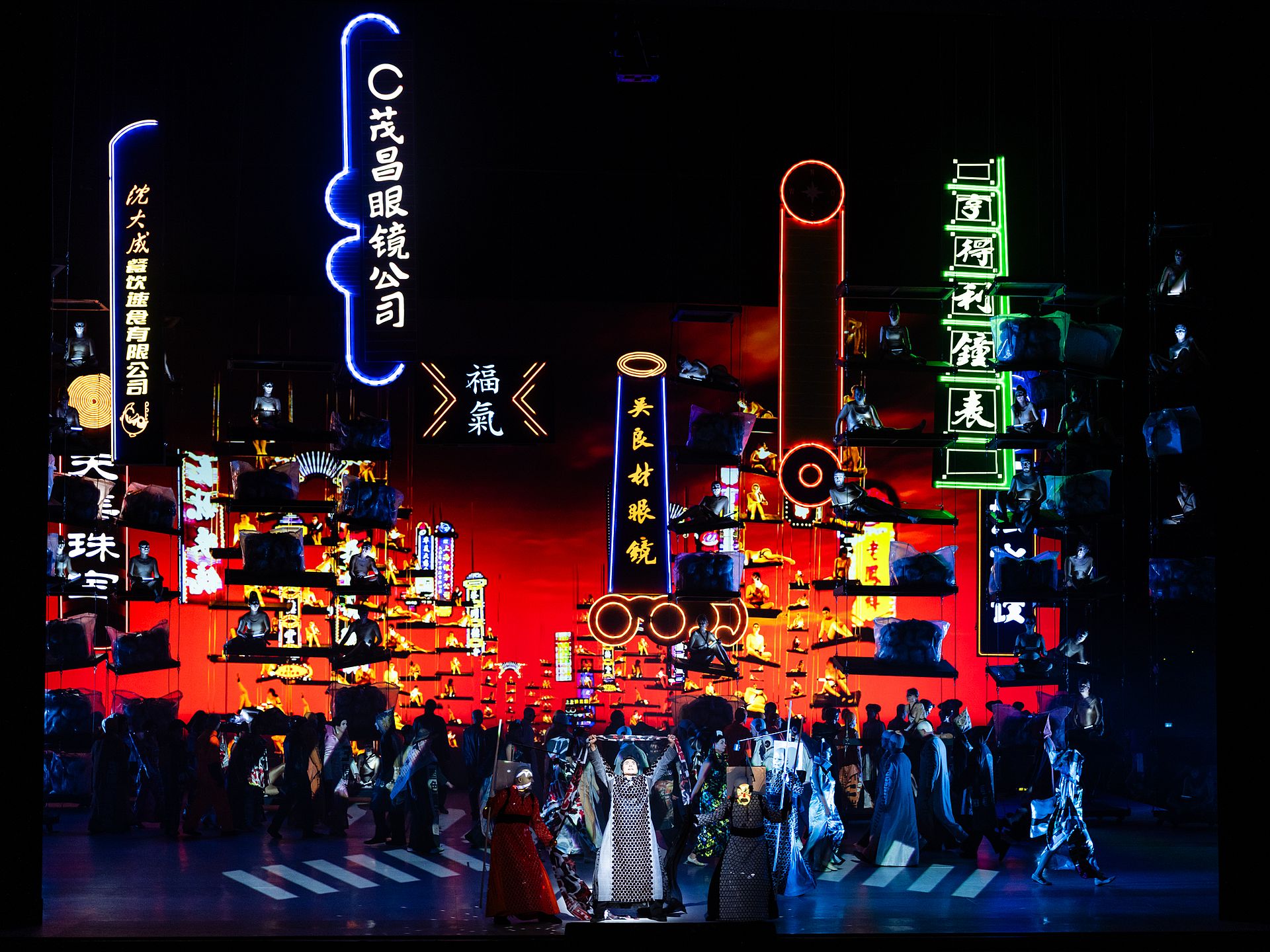 csm_Turandot_2024_c_Geoffroy_Schied__14__c8a8a015a6 - IN München - Das Stadtmagazin Nighttime fashion show on a stage with models in traditional-inspired outfits, neon city signs glowing in the background.
